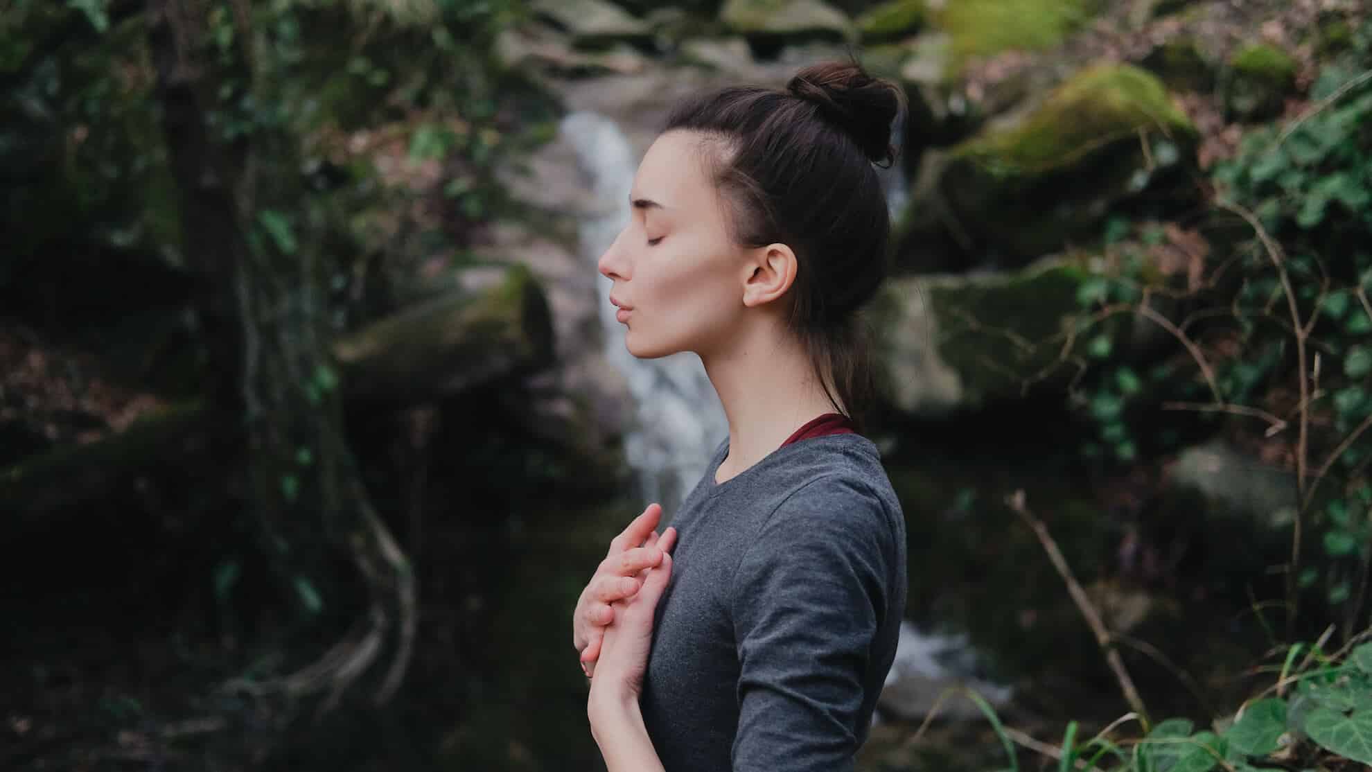 Eine Frau übt Achtsamkeit vor einem ruhigen Wasserfall und findet durch Meditation inneren Frieden.