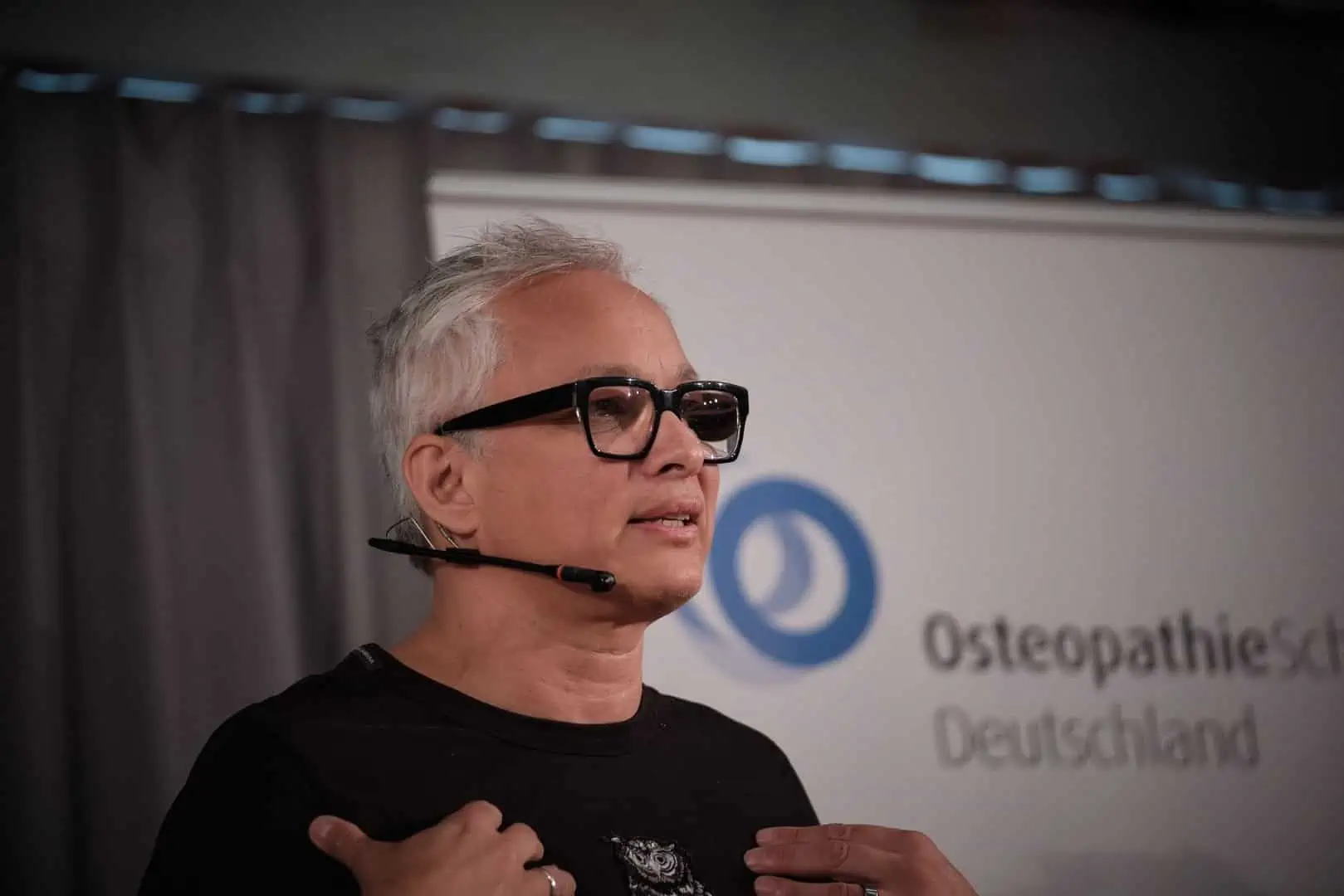 Ein Osteopath mit Brille steht vor einem Schild.