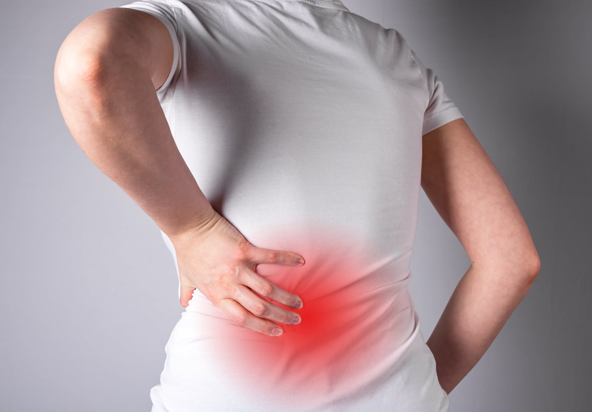 Eine Person im weißen Hemd hält sich vor grauem Hintergrund den unteren Rücken, der durch einen roten Schimmer hervorgehoben ist, der auf chronische Rückenschmerzen im Lendenbereich hinweist.