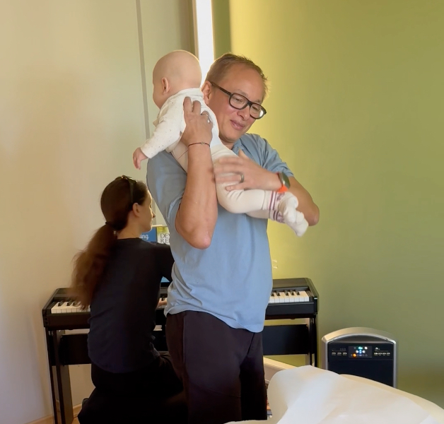 Eine Person mit Brille und hellblauem Hemd hält ein Baby in weißem Outfit. Hinter ihnen spielt eine andere Person mit langem dunklem Haar Klavier und demonstriert damit die heilende Wirkung der Musik. Der Raum hat eine grüne Wand und eine Heizung.