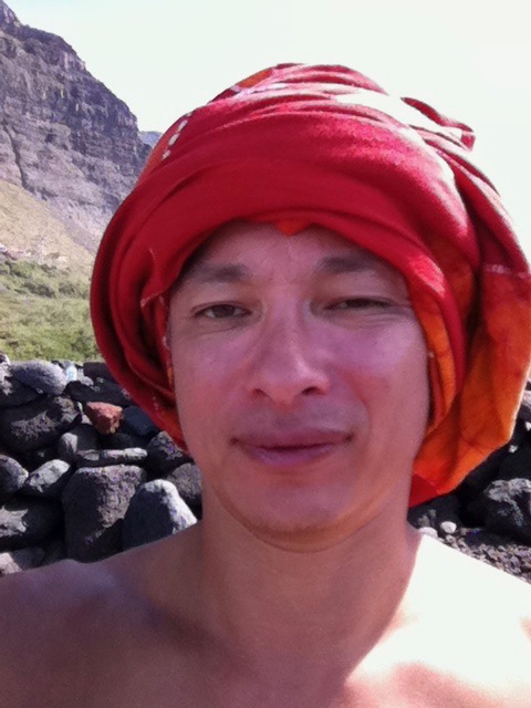 Eine Person mit einem roten Turban ist im Freien mit einer Steinmauer und Bergen im Hintergrund abgebildet. Der Himmel erscheint klar und sonnig und vermittelt ein Gefühl der Ruhe. Die Person, die möglicherweise meditiert, blickt mit einem leichten Lächeln in die Kamera.