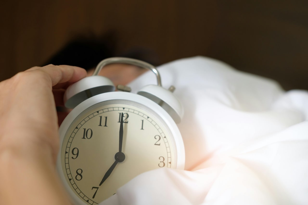 Die Hand einer Person streckt sich aus, um einen kleinen weißen Wecker auszuschalten, der 7:00 Uhr anzeigt. In eine weiße Decke gehüllt, scheint die Person im Bett zu liegen und einen gesunden Schlaf zu genießen, während der leicht verschwommene Hintergrund auf einen ruhigen Morgen hindeutet.