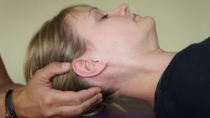 Une personne aux cheveux blonds est allongée sur le dos, les yeux fermés. Une main soutient doucement la tête de la personne sur le côté, peut-être dans le cadre d'un traitement ostéopathique pour un massage ou un ajustement du cou. L'arrière-plan est légèrement flou.