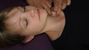 A person with closed eyes lies on their back on a purple surface, wearing a black shirt. A pair of hands gently touches their neck and chest area, employing osteopathische Behandlungsansätze to promote relaxation and well-being.