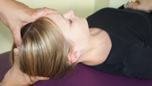 Una persona con i capelli biondi è sdraiata sulla schiena e riceve un massaggio alla testa che ricorda l'osteopathische Behandlungsansätze. Le mani sono delicatamente appoggiate ai lati della testa. La persona indossa una maglietta nera e si appoggia su una superficie viola, godendosi questo metodo di rilassamento olistico.