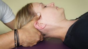 Person liegt auf dem Rücken, während die Hände einer anderen Person ihren Kopf stützen, eine in osteopathischen Behandlungsansätzen übliche Geste. Die stützende Person trägt ein schwarzes Armband. Der Hintergrund ist neutral und beide Personen haben helle Hauttöne.