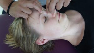A person lying down receives a facial massage, incorporating osteopathische Behandlungsansätze. Hands gently apply pressure to the forehead and nose bridge. With closed eyes, the person appears relaxed, their light brown hair contrasting with a black top.
