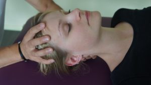 Una persona è sdraiata su una superficie viola, con gli occhi chiusi, e si gode un massaggio alla testa con mani delicate che applicano osteopathische Behandlungsansätze. Indossando un top nero, si crogiolano nel relax mentre lo sfondo sfuma dolcemente.