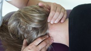 Osoba z długimi blond włosami leży twarzą w dół na powierzchni, podczas gdy inna osoba delikatnie stosuje osteopathische Behandlungsansätze, używając obu rąk, aby ukoić szyję. Osoba otrzymująca tę opiekę ma na sobie czarną koszulę, całkowicie zrelaksowana pod fachowym dotykiem praktyka.