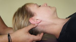 Une personne touche doucement le cou d'une autre personne, allongée avec la tête tournée sur le côté, en appliquant des techniques de traitement ostéopathiques. L'accent est mis sur le contact entre la main et le cou, facilitant un sentiment de calme et favorisant une mise à jour du bien-être.