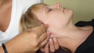 Osoba z krótkimi blond włosami leży na plecach, z zamkniętymi oczami, podczas gdy inna osoba delikatnie masuje okolicę ucha, stosując osteopathische Behandlungsansätze. Scena przywołuje relaksujący, terapeutyczny zabieg skupiający się na technikach drenażu mózgu.