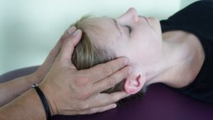 Eine Person mit heller Haut und schwarzem Hemd liegt friedlich mit geschlossenen Augen da und erhält eine sanfte Kopfmassage. Dieser osteopathische Behandlungsansatz scheint die Entwässerung des Gehirns zu erleichtern. Die Hände werden sanft auf beiden Seiten des Kopfes vor einem unscharfen Hintergrund positioniert.